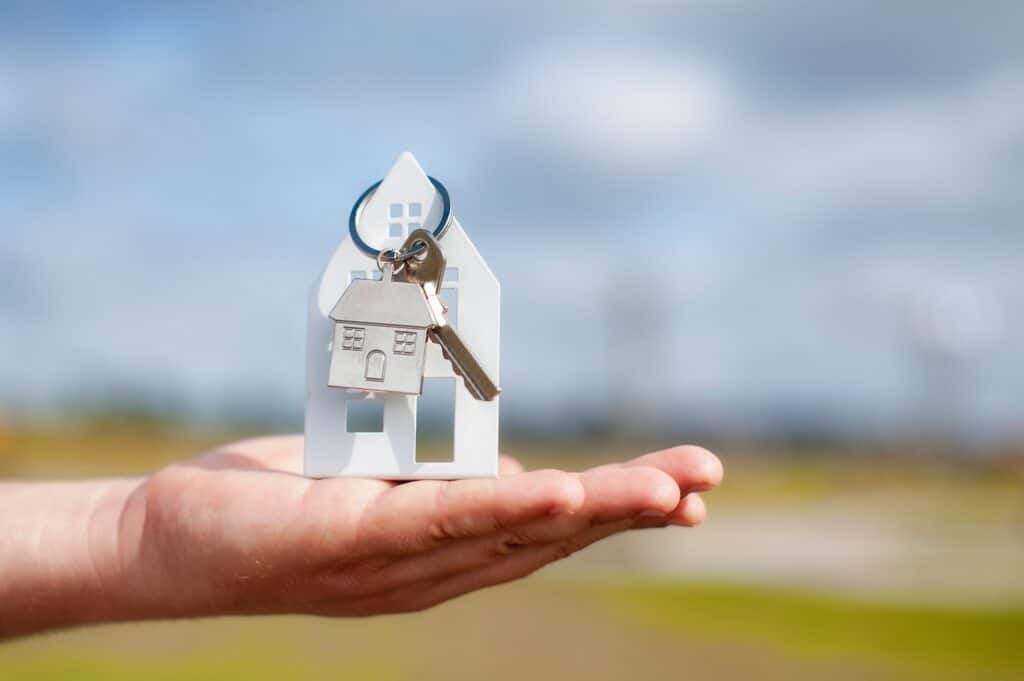 une main tient une maison en papier sur laquelle il y a un porte-clé avec une clé et une maison blanche