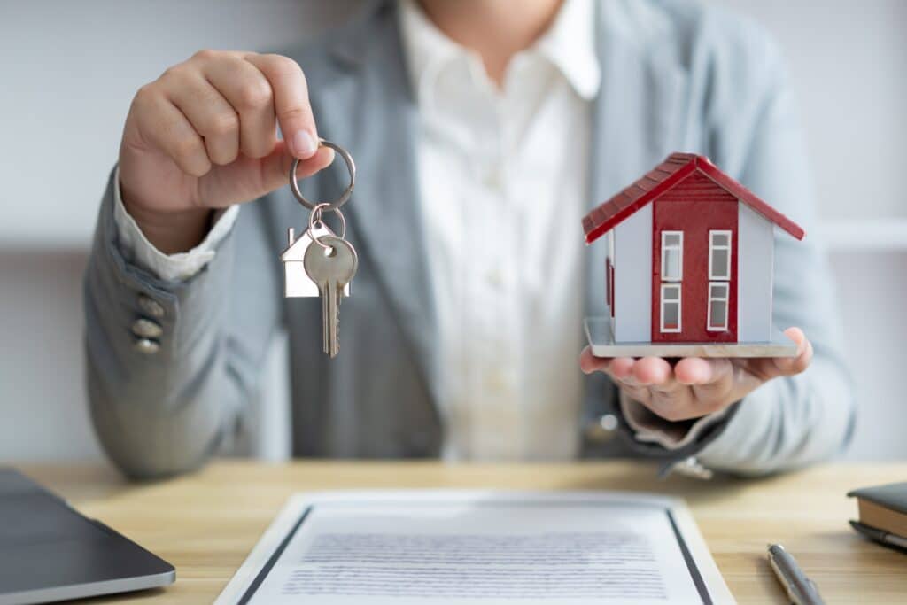 Une personne tenant dans une main les clés d'une maison et dans l'autre la maquette d'une maison. devant lui est posé un document sur une table en bois.