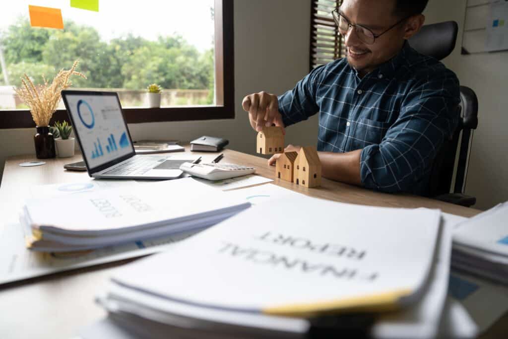 Un homme souriant habillé de chemise à carreaux et portant des lunettes assis devant son bureau sur lequel sont posés des documents, une calculatrice et un ordinateur portable. Il aligne des maisons en bois miniatures sur le bureau.
